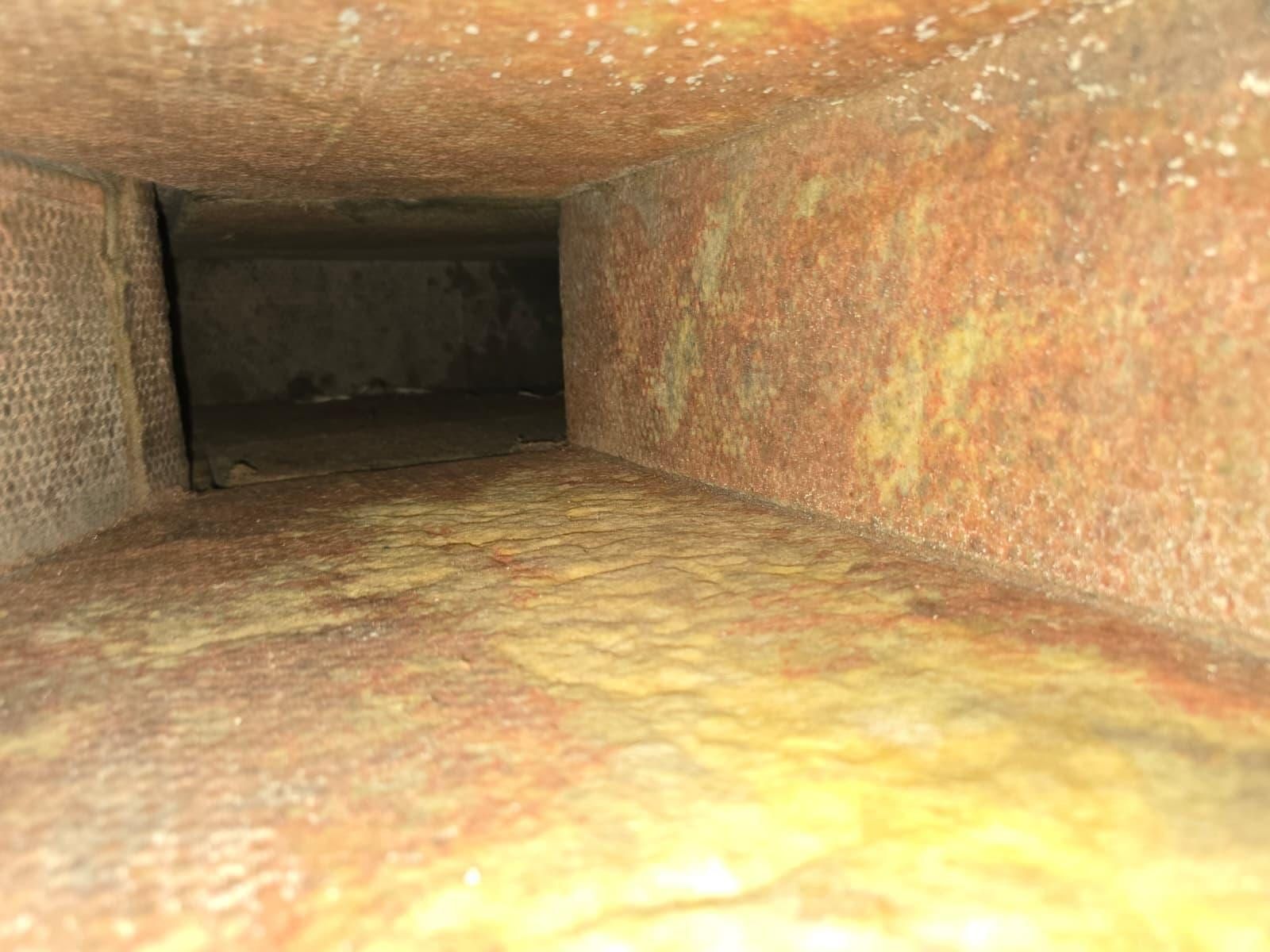 El interior de un pleno de ducto de panel de fibra (duct board), con las paredes muy manchadas de marrón anaranjado por los años