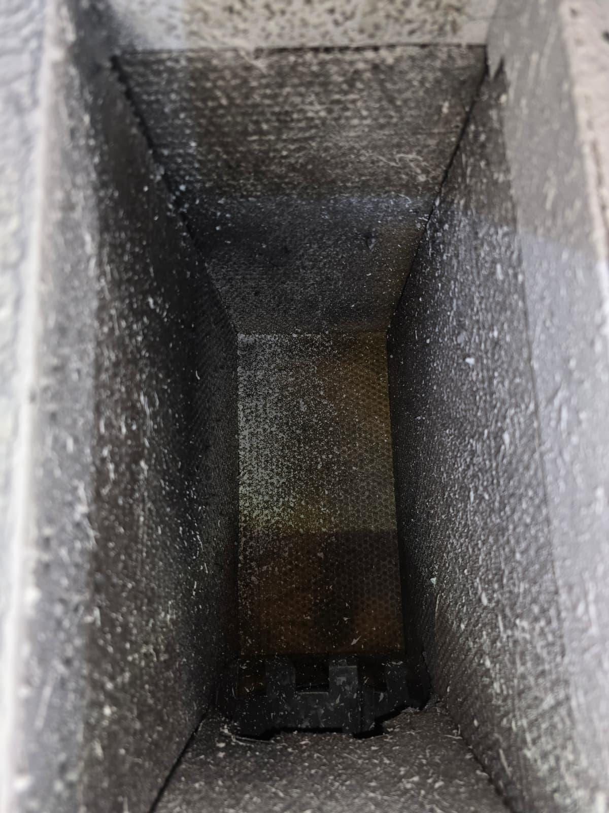 El interior de un ducto rectangular estrecho, cubierto de una gruesa capa de polvo gris con textura, que se estrecha hacia una abertura oscura con junta